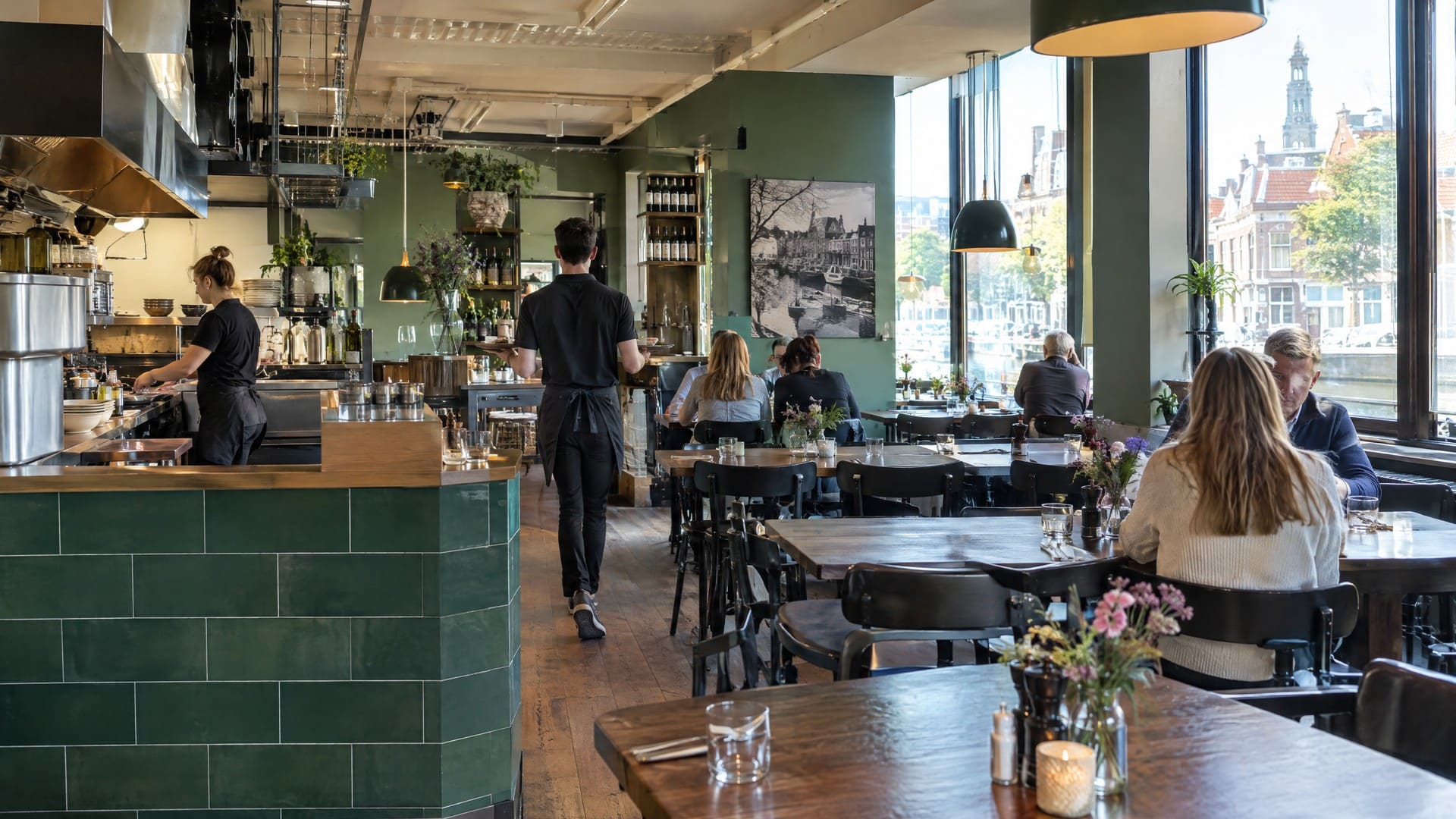 Bistro met groene tegels, houten tafels en een open keuken, met uitzicht op het water